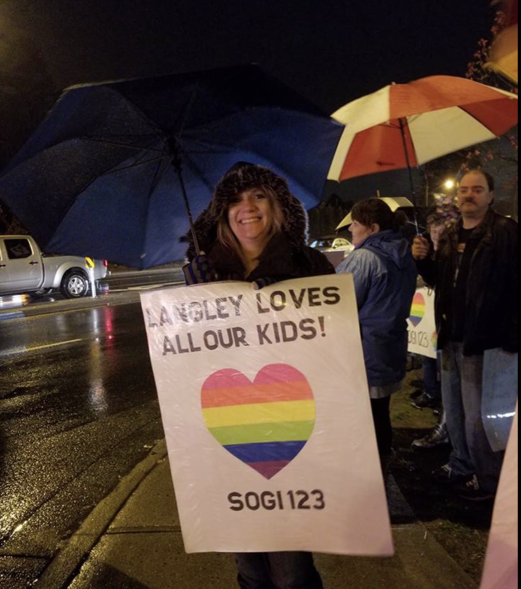 A smiling woman under an umbrella on a rainy night holds a sign reading “Langley loves all our kids! SOGI 123” with a rainbow heart, as other supporters with signs stand beside her
